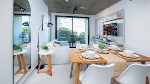 Uma área de refeições moderna com mesa de madeira posta para quatro pessoas, cadeiras brancas e uma grande janela com vista para o exterior.