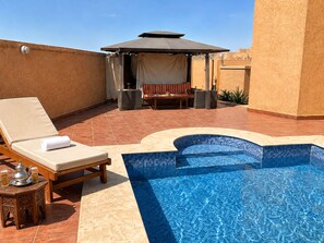 A rooftop pool area with a lounge chair, a small table, and a covered seating area.