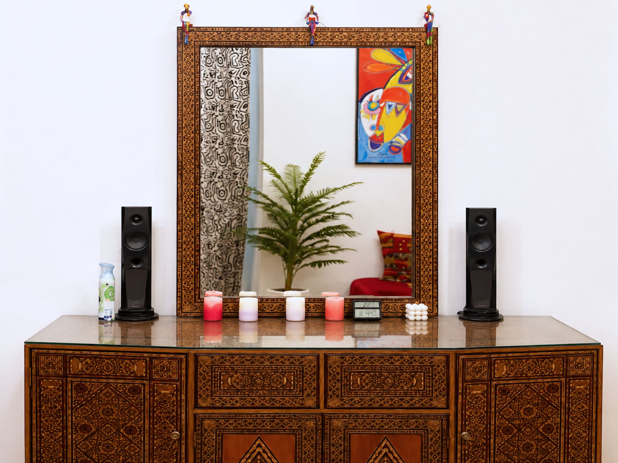 Un mueble de madera con intrincados grabados, un espejo con marco decorativo, una planta en maceta y dos altavoces a cada lado.