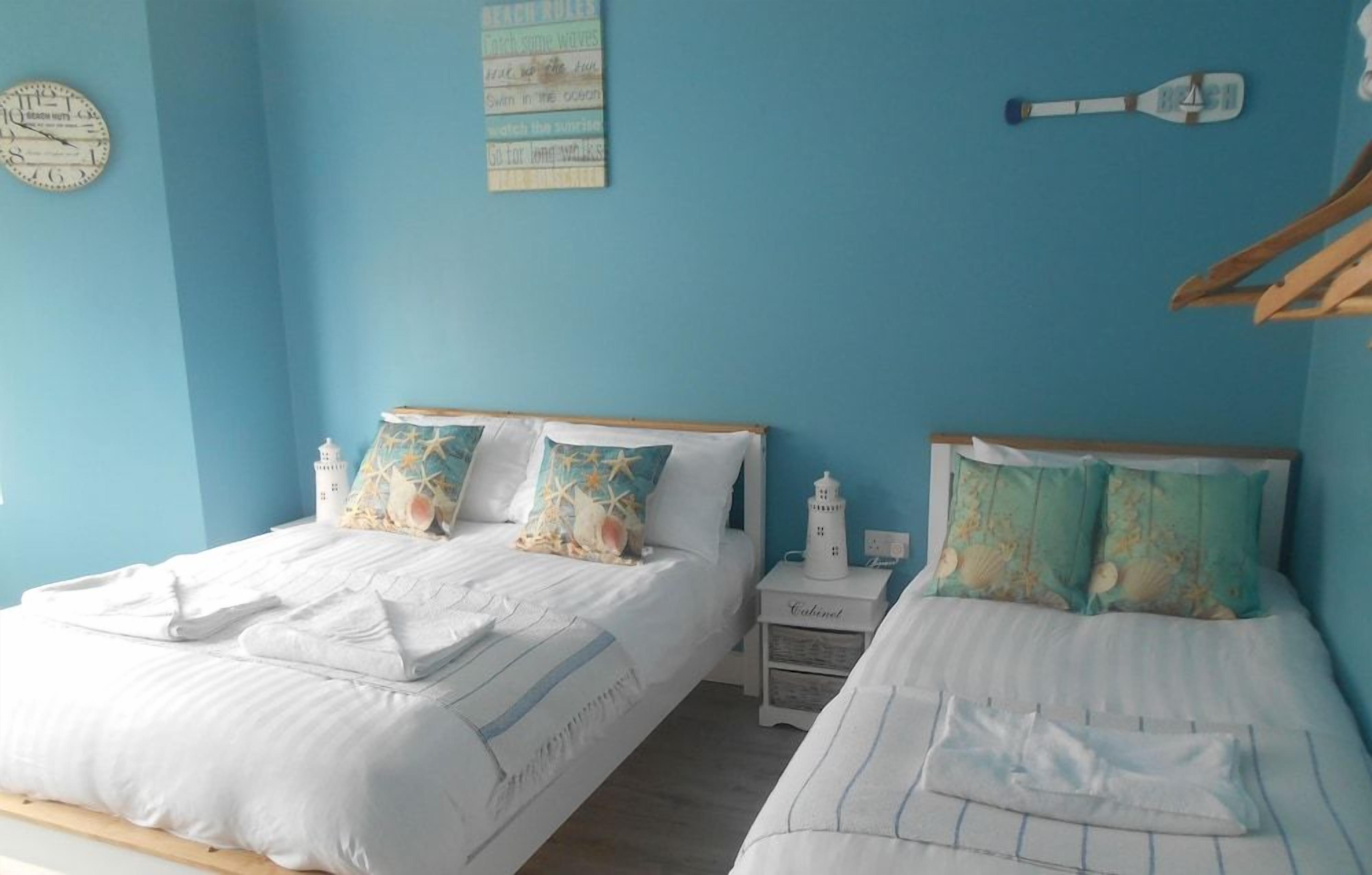 A room with two beds, each with white bedding and decorative pillows, a small nightstand with a lamp, and a wall clock.