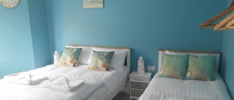A room with two beds, each with white bedding and decorative pillows, a small nightstand with a lamp, and a wall clock.