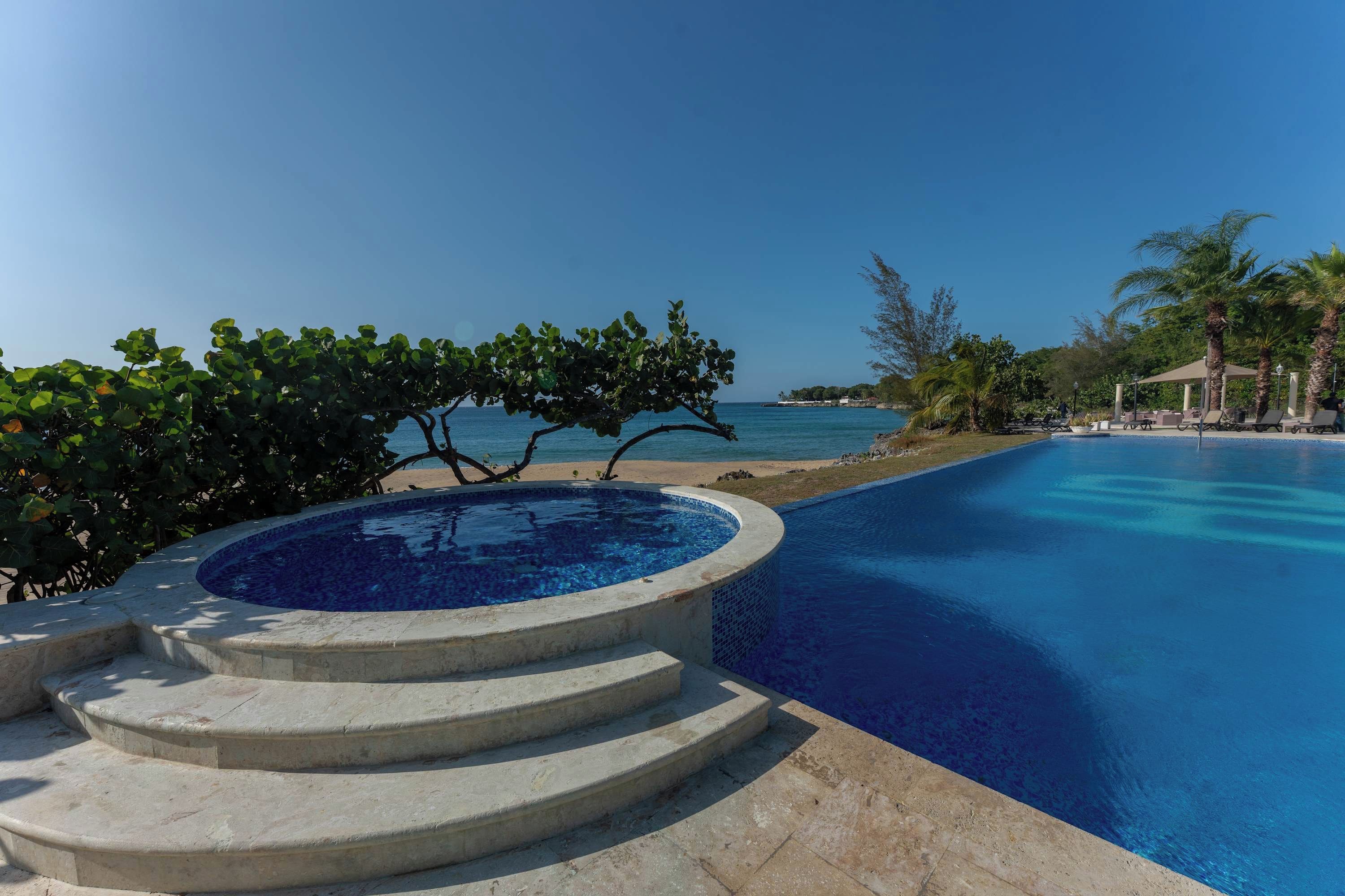 Une piscine d’un bleu éclatant, avec des marches permettant d’y descendre, entourée de palmiers et offrant une vue sur la mer.