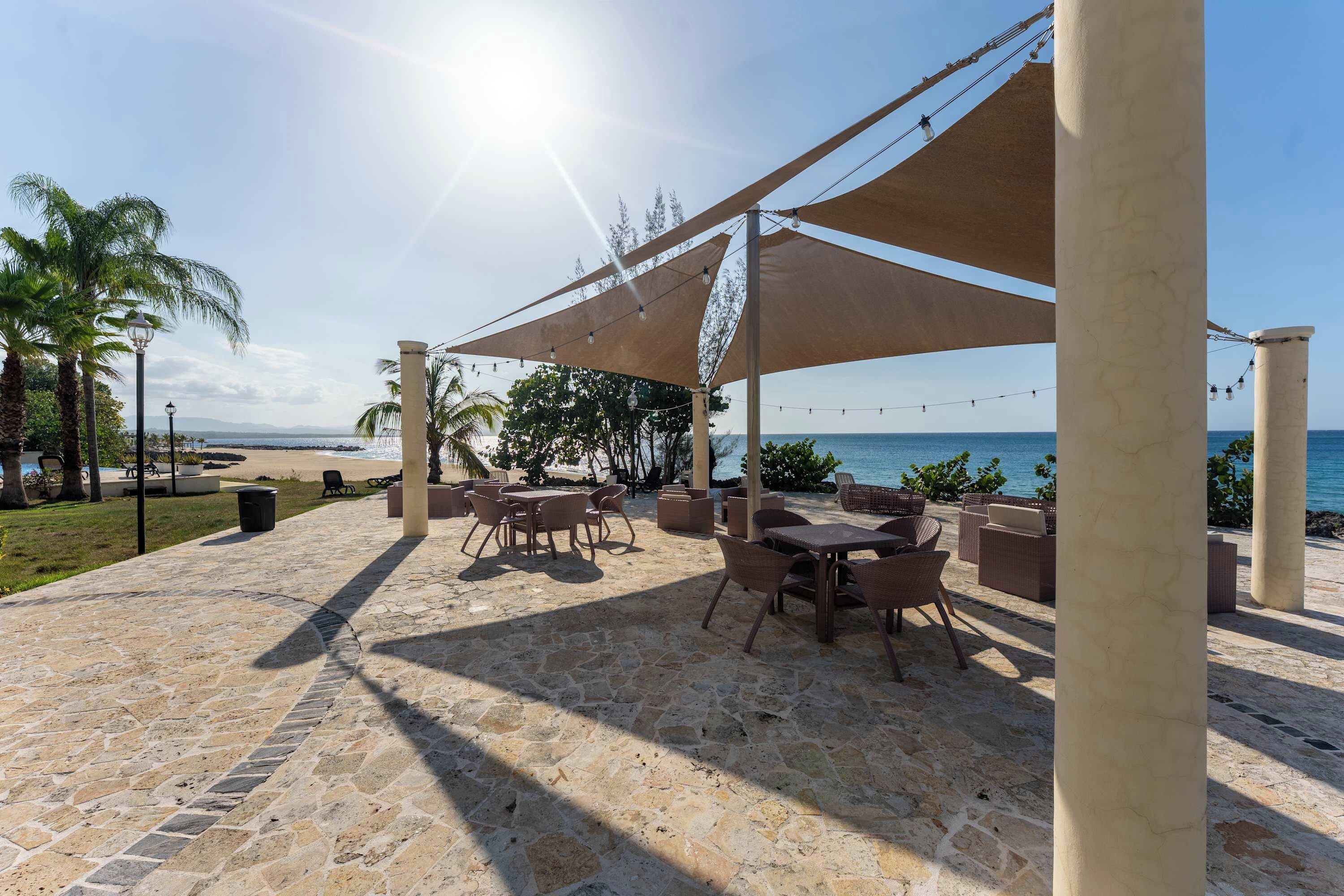An outdoor patio with stone flooring, tables, chairs, and a canopy providing shade.