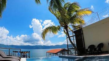 A pool area with a view of the sea and mountains, a hot tub, lounge chairs, and a covered seating area.