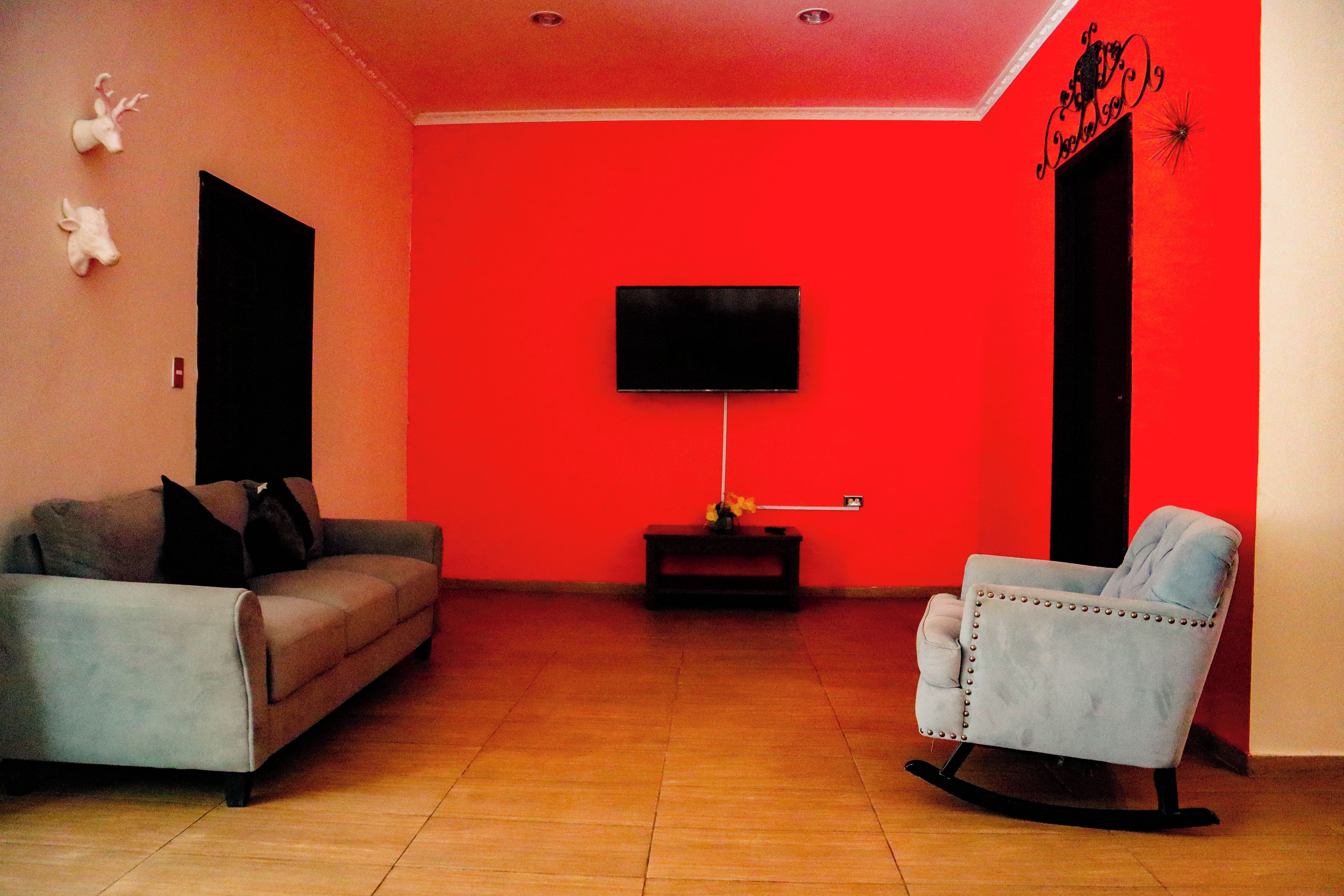 Una sala de estar con una pared roja, un sofá gris, una mecedora, un televisor de pantalla plana montado en la pared y una mesita con un jarrón.