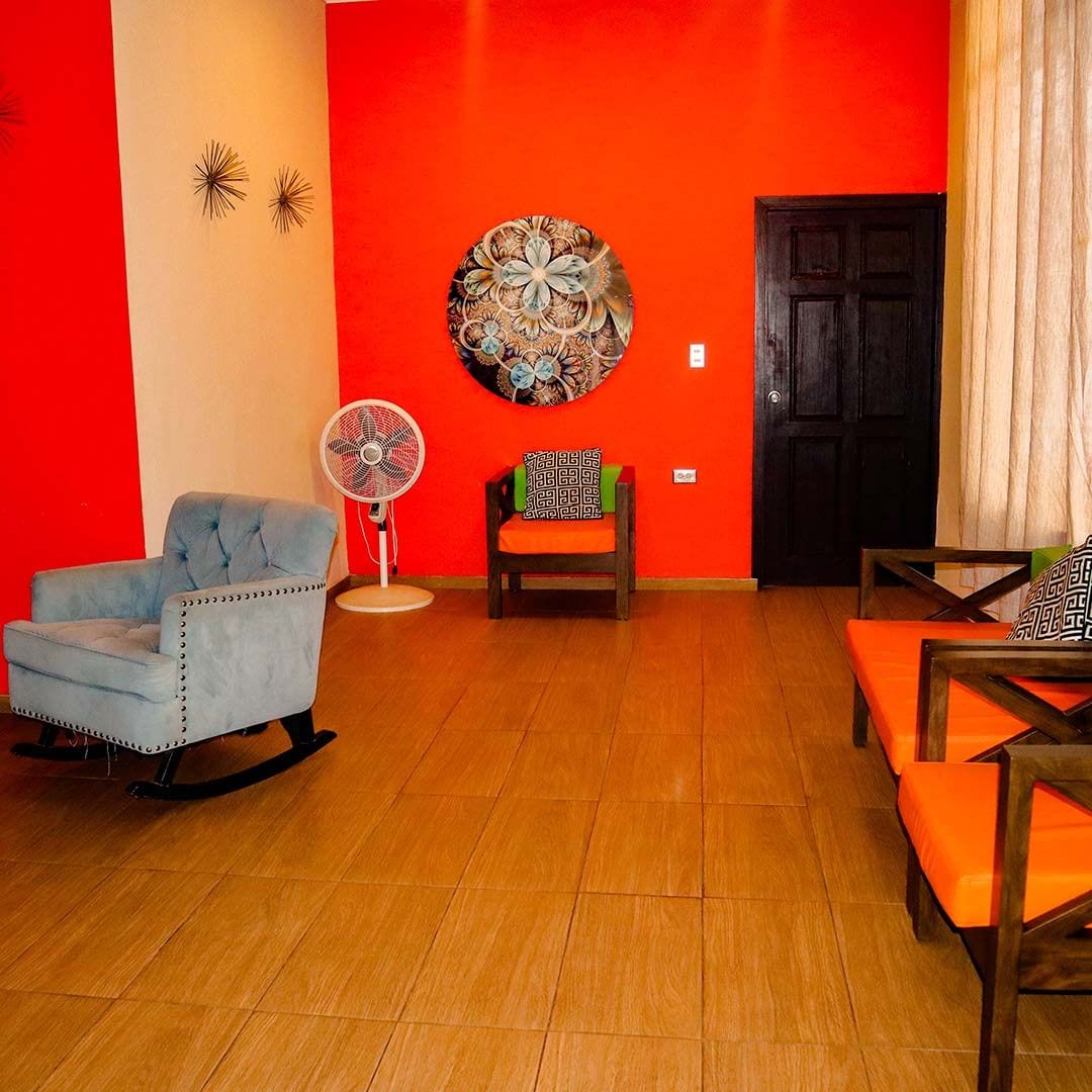 Una sala de estar con una mecedora azul, un ventilador, una silla de madera con cojín y un sofá con cojines naranjas.