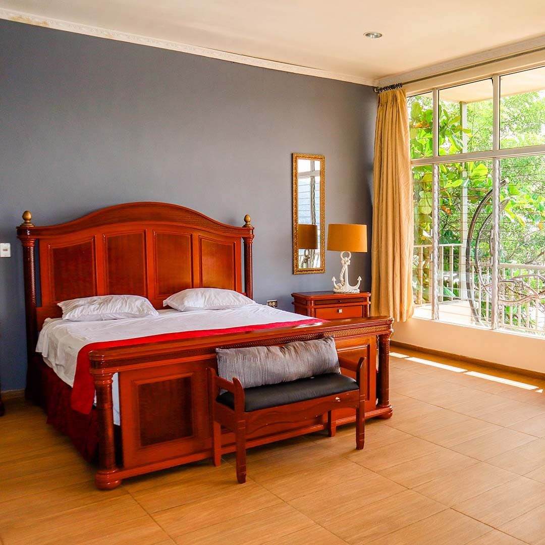 A bedroom with a large bed, a bedside table with a lamp, a mirror, and a window with a view of greenery.