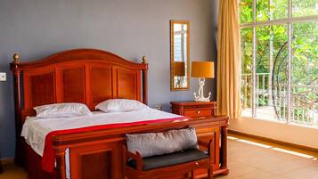 A bedroom with a large bed, a bedside table with a lamp, a mirror, and a window with a view of greenery.