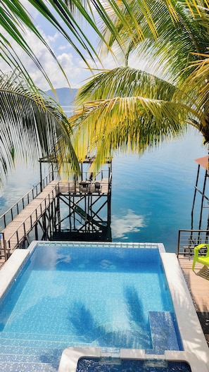 Une piscine d’un bleu éclatant, avec une terrasse en bois et des palmiers, surplombant une étendue d’eau calme.
