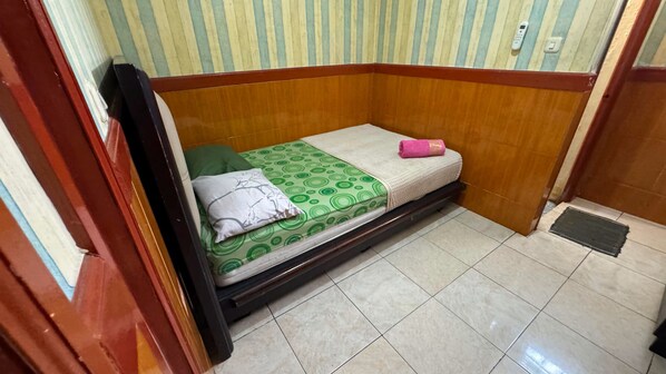 A small, single-bed room with a wooden headboard, a green patterned bedspread, a white pillow, and a pink towel.