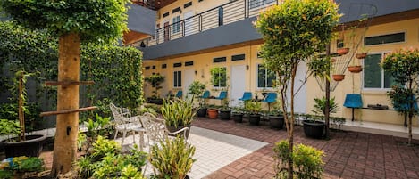 Un patio con plantas en macetas, mesas al aire libre y un edificio con varias ventanas y balcones.