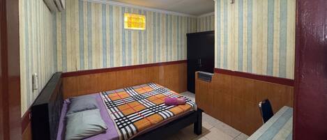 A small, single-bed room with striped wallpaper, a wooden headboard, a bedside table, and a ceiling light.
