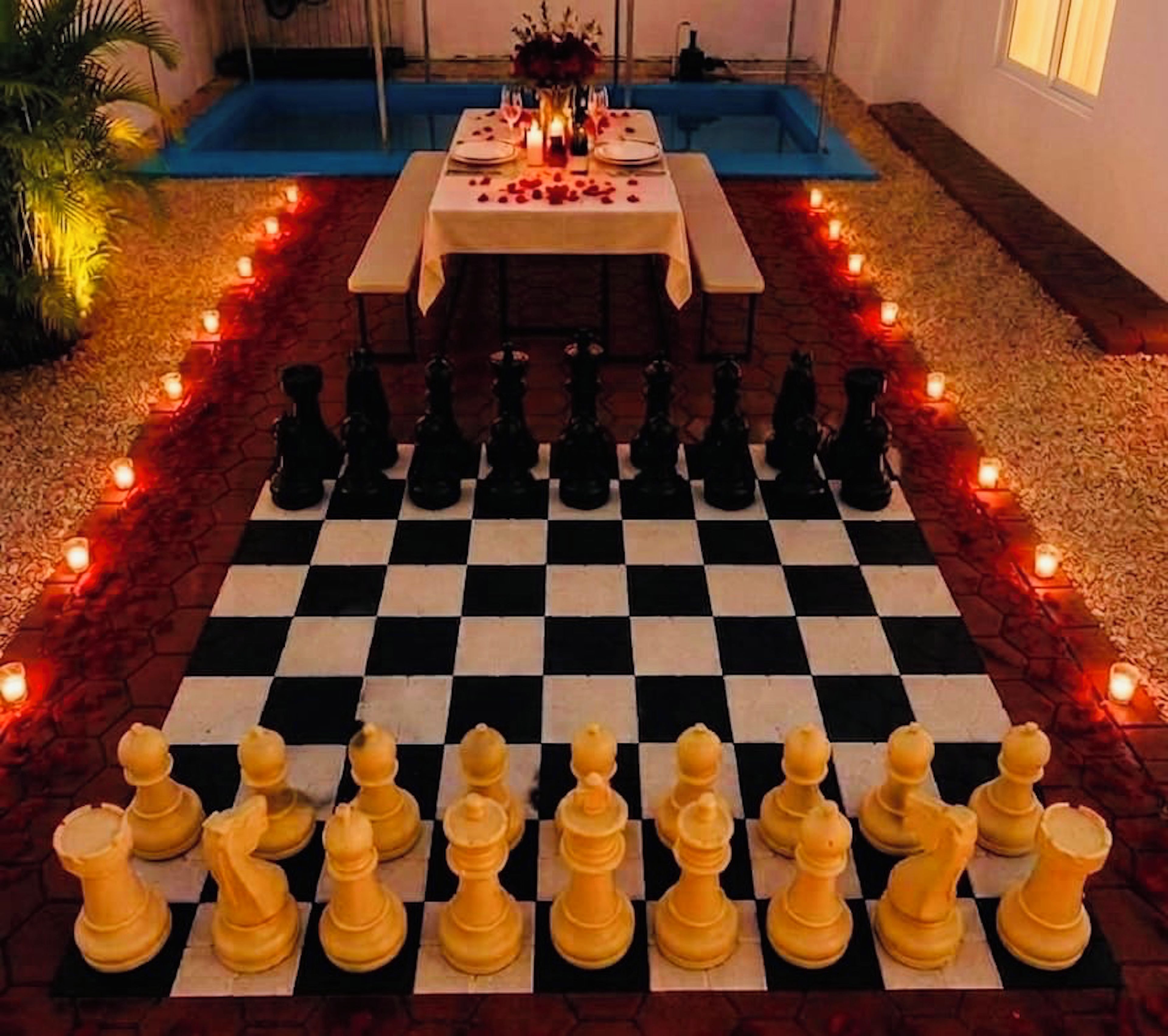 A checkered chessboard with pieces arranged for a game, surrounded by candles and a table with a floral centerpiece.