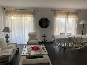Ein Wohnzimmer mit einer Couch, einem Esstisch mit Stühlen, einem kleinen Tisch mit Blumenvase und einer Lampe.