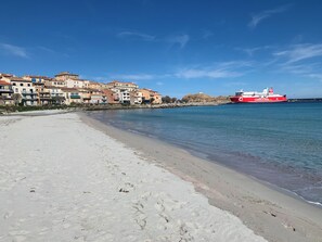 Ein Strand mit einem roten Schiffen in der Ferne, Gebäuden entlang der Küste und blauem Himmel.