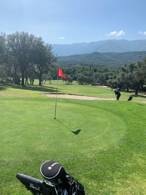 Ein Golfplatz mit einer roten Fahne, Bäumen und Bergen im Hintergrund.