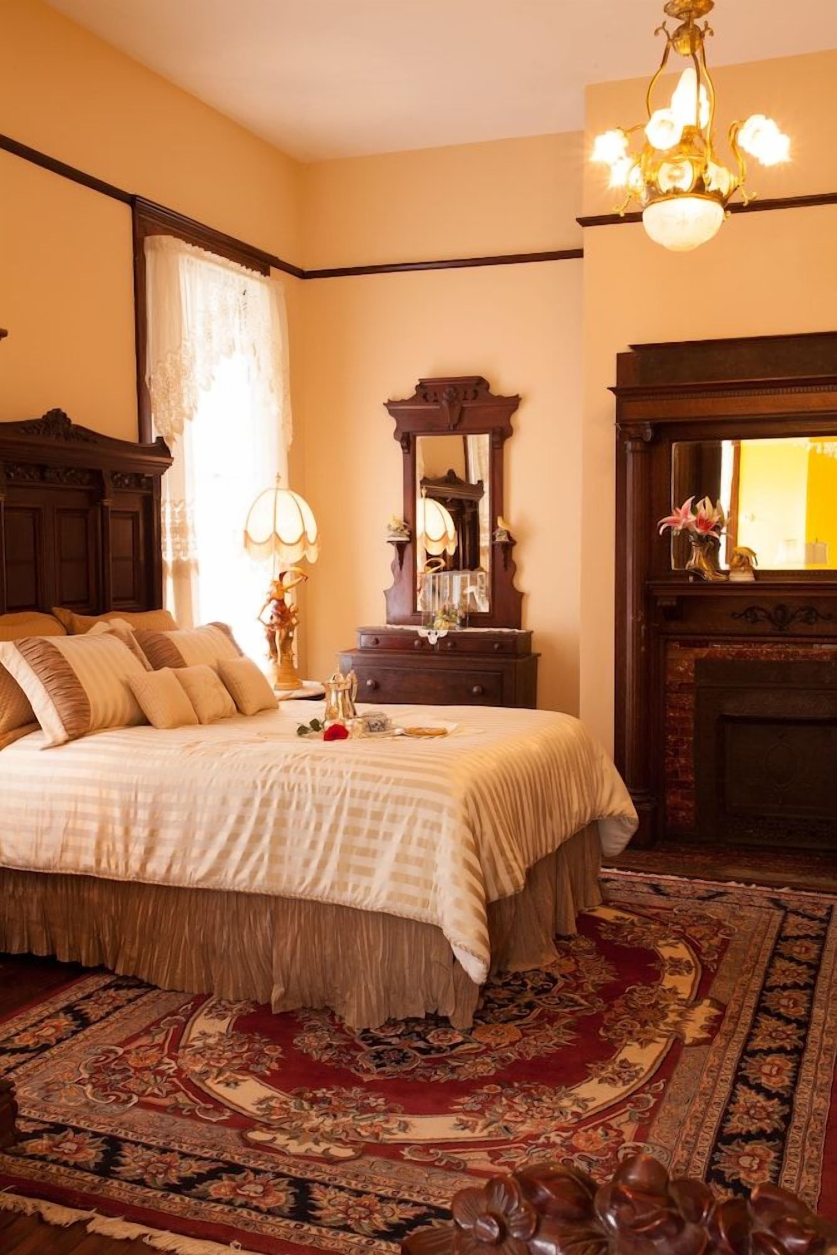 A traditional bedroom with a large bed, a wooden dresser, a mirror, and a fireplace.