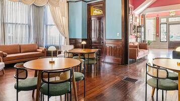 A room with a brown leather sofa, round wooden tables with chairs, and a large window with curtains.