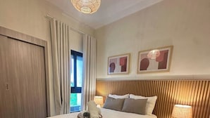 Un dormitorio con cama, lámparas de noche, una bandeja decorativa, cabecero de madera y una ventana con cortinas.