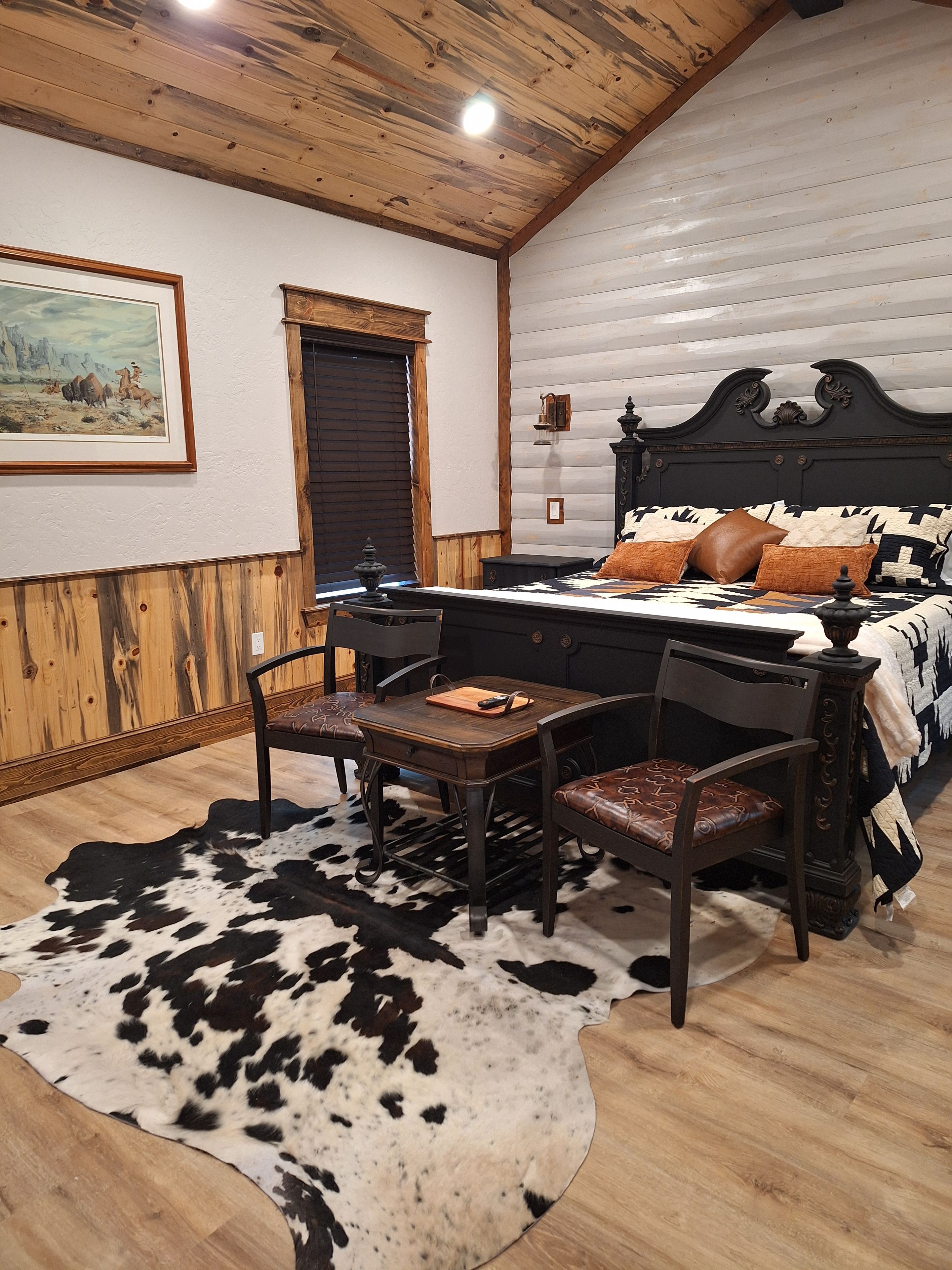 A bedroom with a wooden ceiling, a bed with patterned bedding, a cowhide rug, and a dining table with chairs.