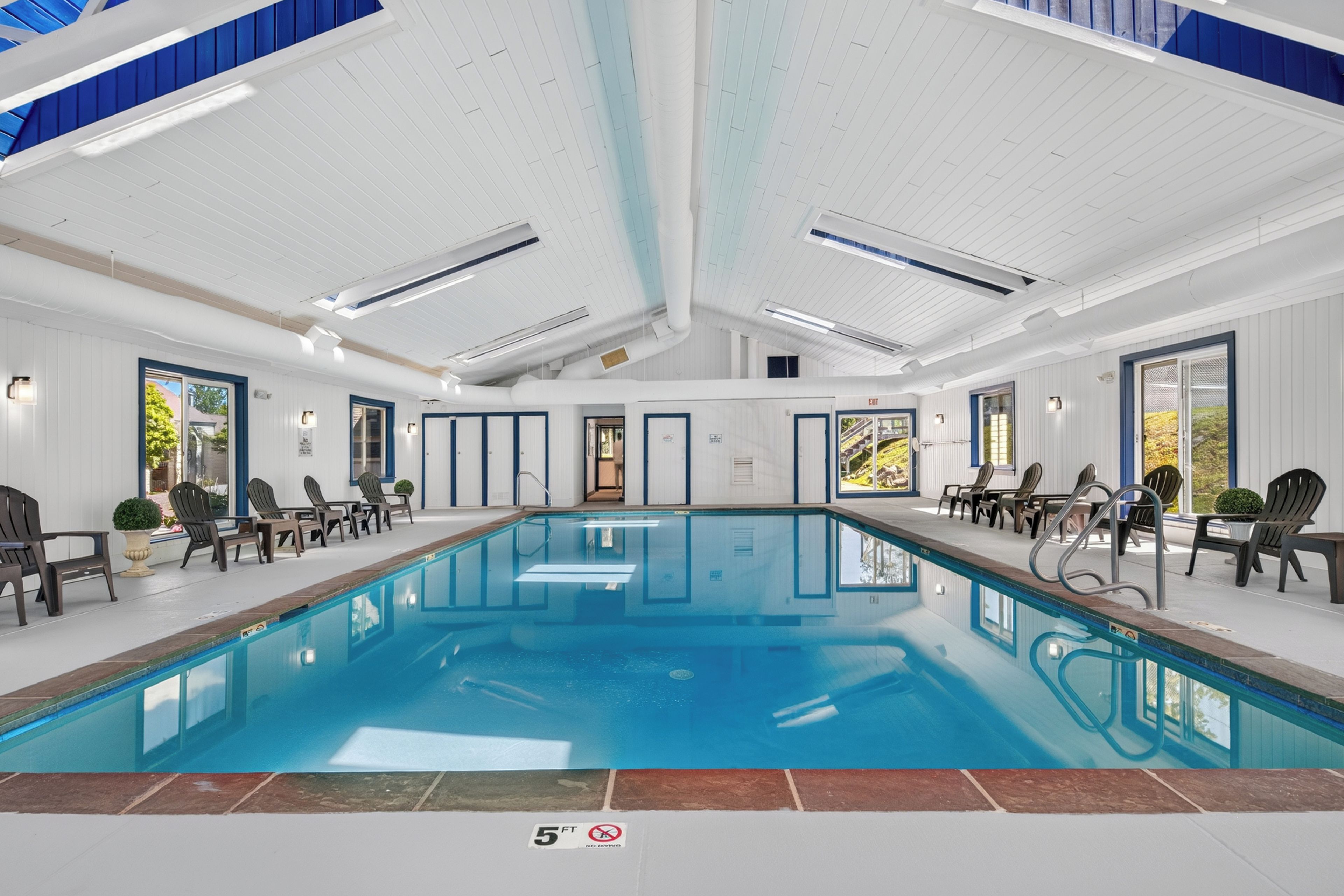 A large indoor swimming pool with clear blue water, surrounded by lounge chairs and a high ceiling with skylights.