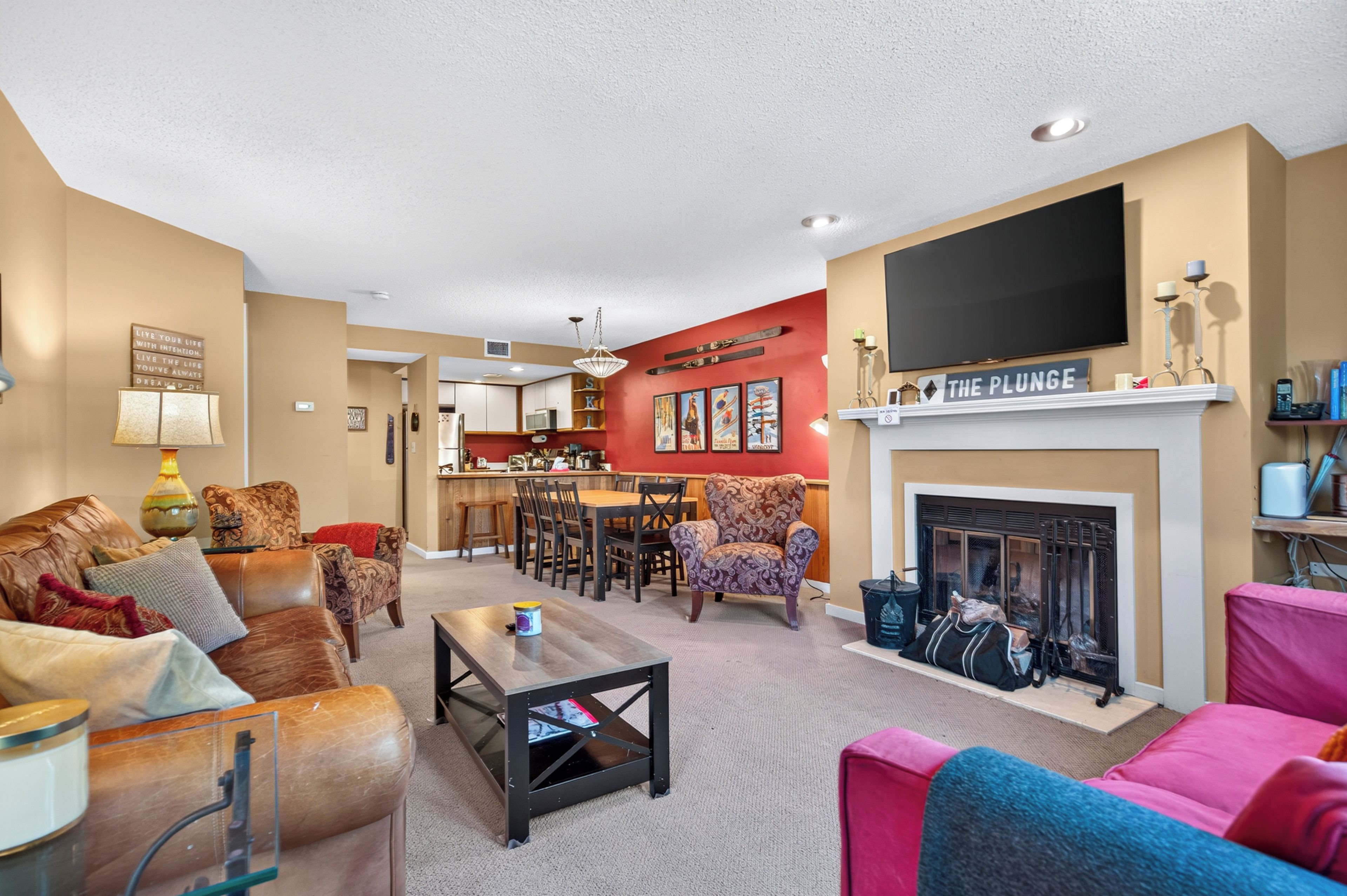 A living room with a fireplace, a television mounted above it, and a kitchen in the background.