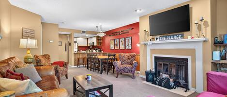 A living room with a fireplace, a television mounted above it, and a kitchen in the background.