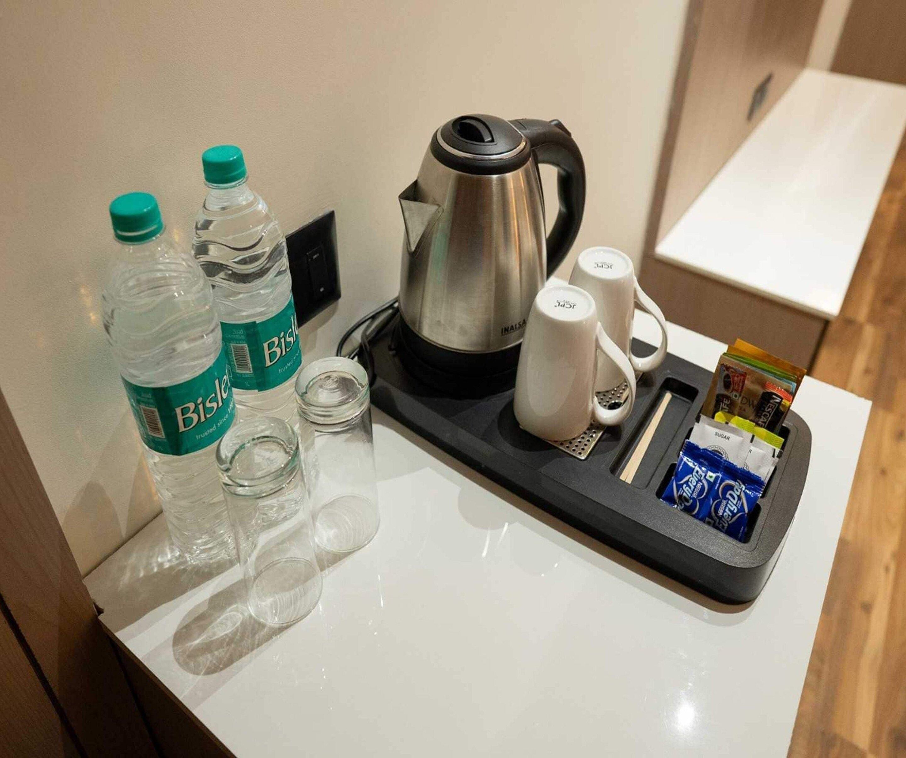 A hotel room's minibar with Bisleri water bottles, a kettle, coffee mugs, and snacks.