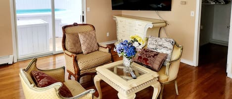 Un salon avec un téléviseur fixé au mur, une table basse en verre ornée d’un vase de fleurs et deux fauteuils avec des coussins.
