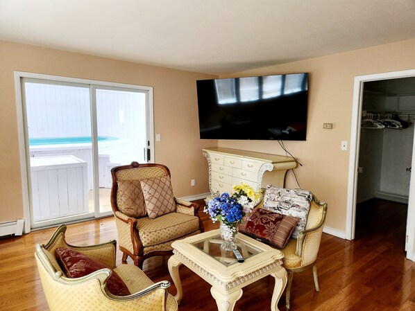 A living room with a television mounted on the wall, a glass coffee table with a vase of flowers, and two armchairs with cushions.