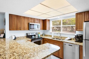 A modern kitchen with granite countertops, wooden cabinets, a built-in microwave, a stainless steel refrigerator, and a window with blinds. - First Floor Cottage at Hidden Dunes, Near Tennis Courts (Miramar Beach)