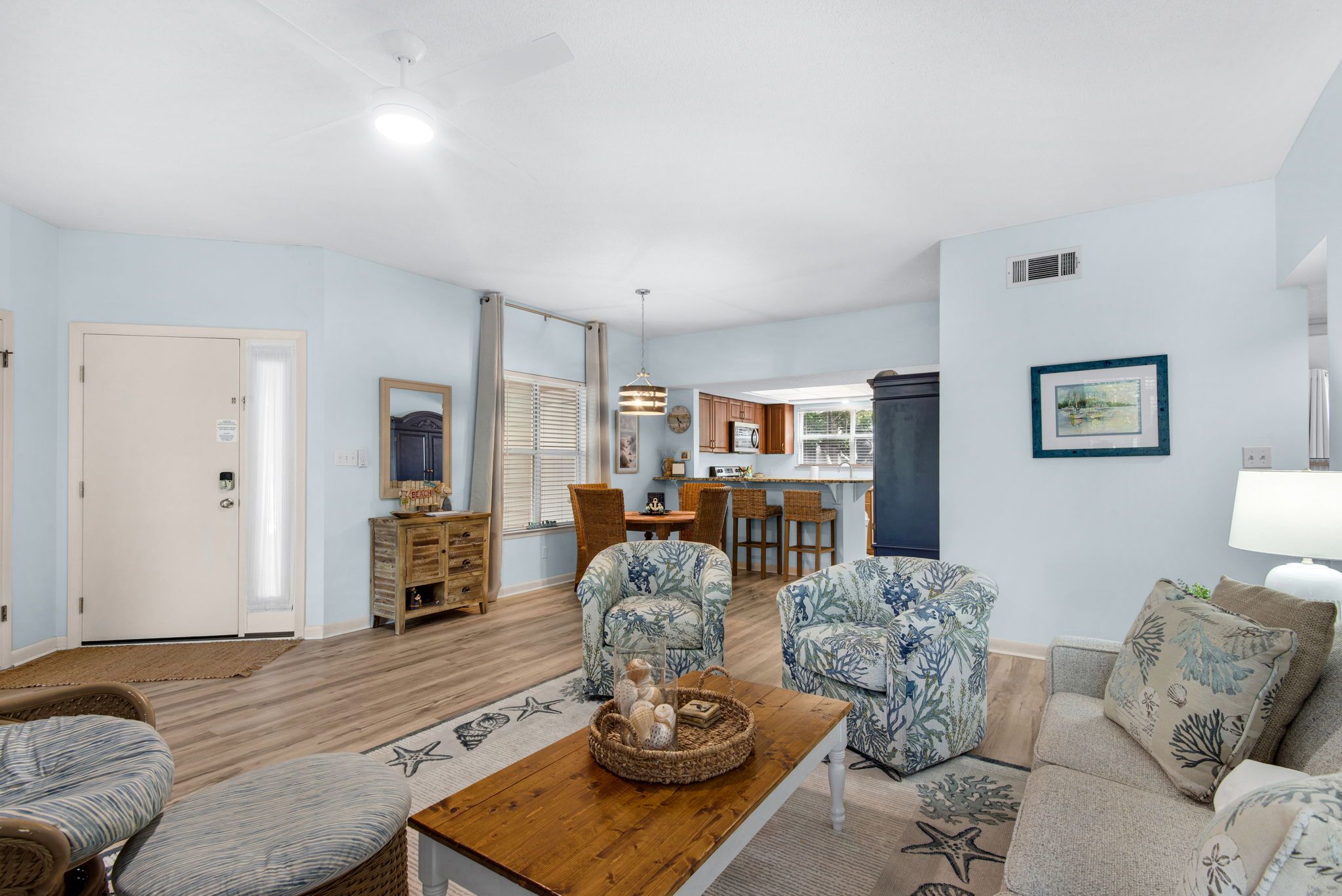 A spacious living room with a wooden coffee table, two patterned armchairs, a sofa, and a kitchen area in the background.
