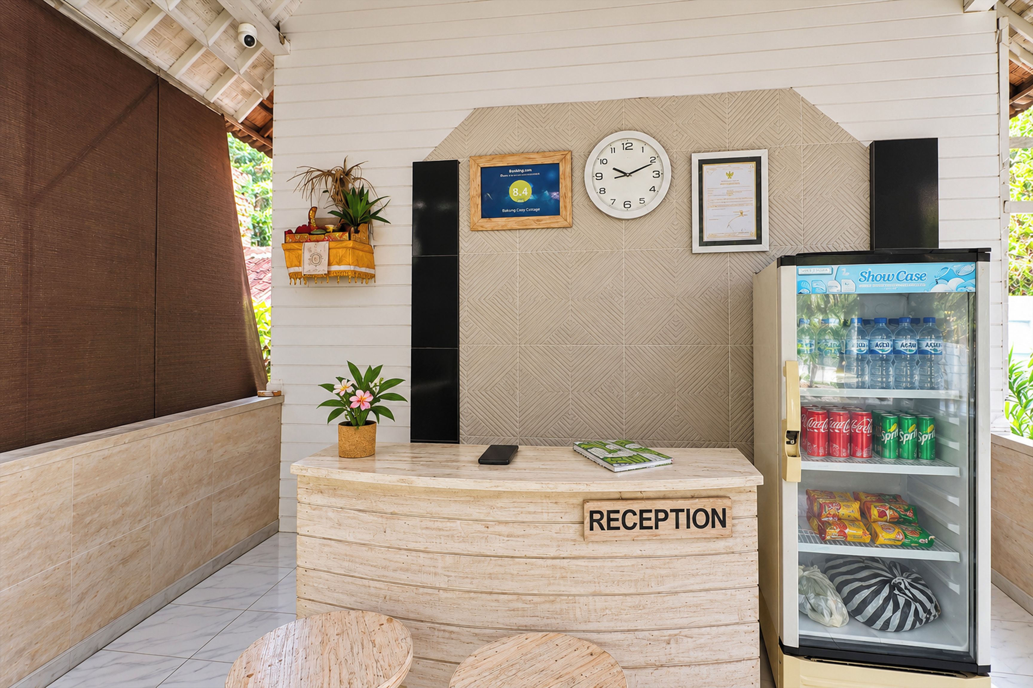 Un área de recepción con un mostrador de madera, un reloj, diplomas enmarcados, un refrigerador con bebidas y una planta en maceta.