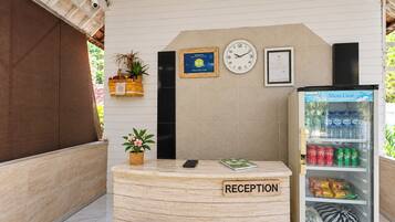 Un área de recepción con un mostrador de madera, un reloj, diplomas enmarcados, un refrigerador con bebidas y una planta en maceta.