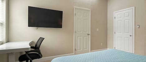 Une chambre à coucher avec un lit, un bureau et une chaise. Il y a deux portes et un téléviseur fixé au mur.