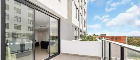 En moderne bygning med store glasvinduer, en balkon med gelænder og udsigt til andre bygninger og grønt.