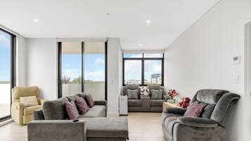 A modern living room with a sofa, armchairs, and a coffee table. There are large windows providing a view of the city.