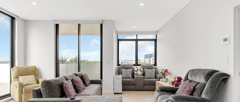 A modern living room with a sofa, armchairs, and a coffee table. There are large windows providing a view of the city.