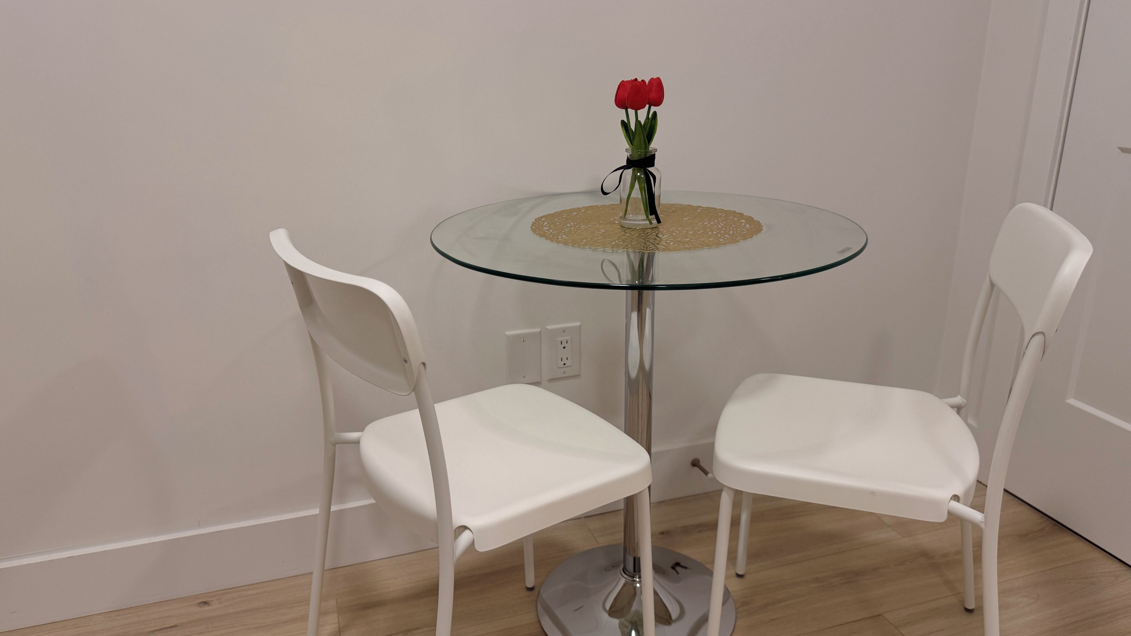 A small round glass table with a vase of red flowers, flanked by two white chairs.