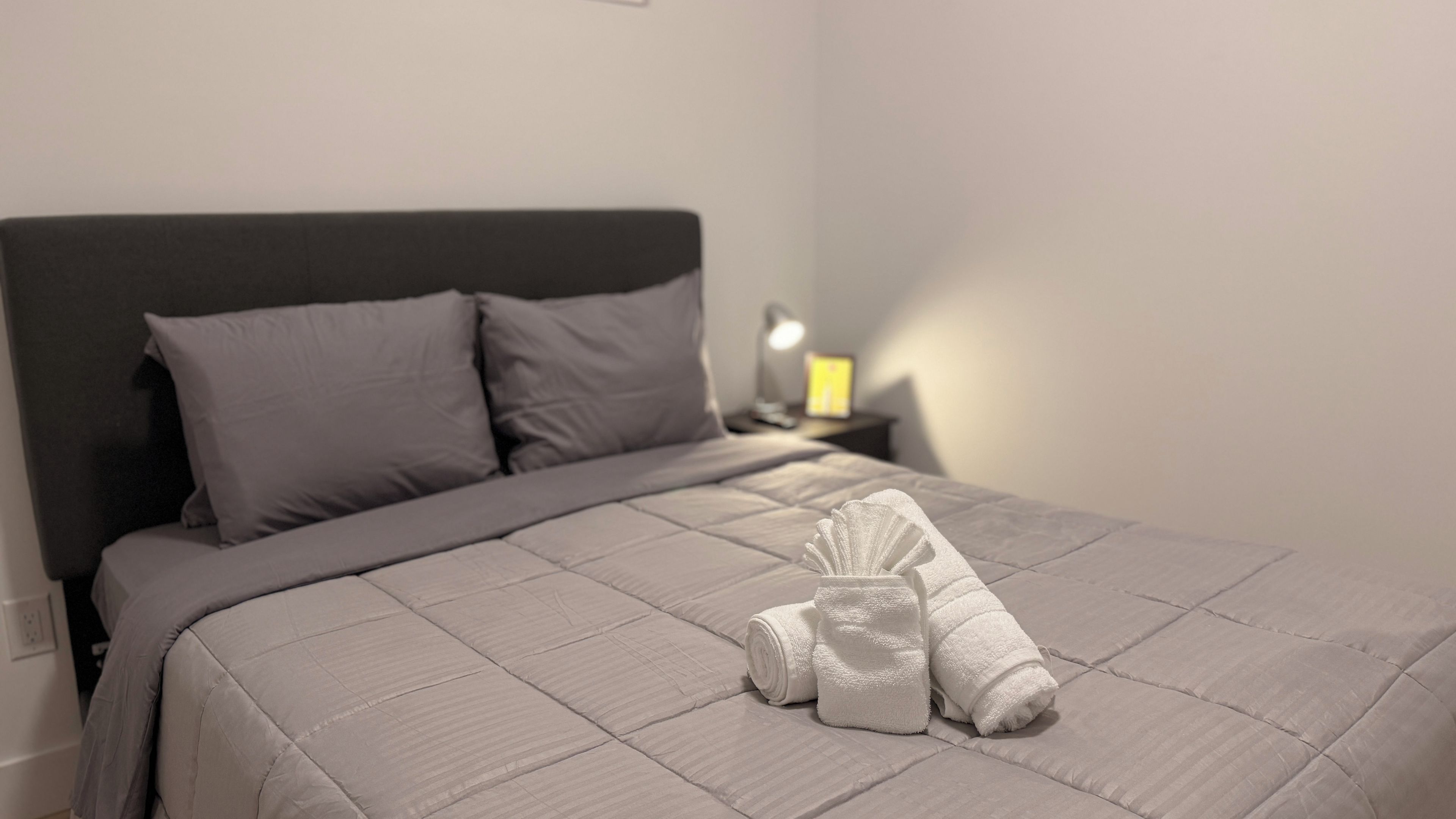 Una cama bien hecha con edredón y almohadas grises, una toalla doblada sobre la cama y una lámpara de noche en una mesita.