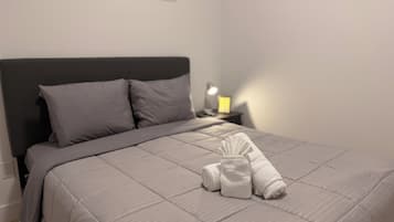 Una cama bien hecha con edredón y almohadas grises, una toalla doblada sobre la cama y una lámpara de noche en una mesita.