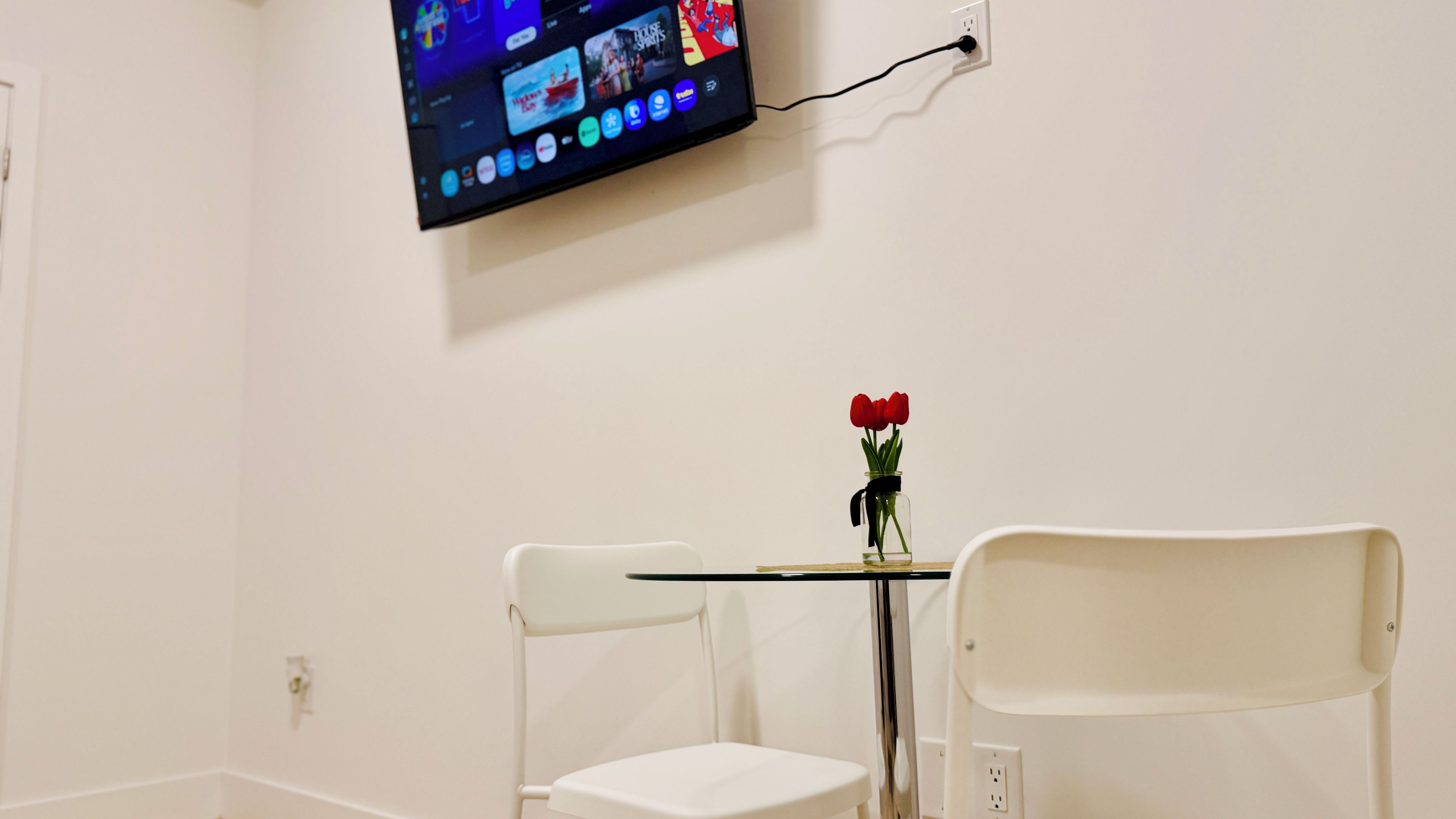 A room with a television mounted on the wall, a small table with a vase of flowers, and two white chairs.