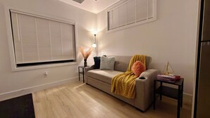 Een woonkamer met een bank, een bijzettafeltje, een lamp en een raam met zonwering.
