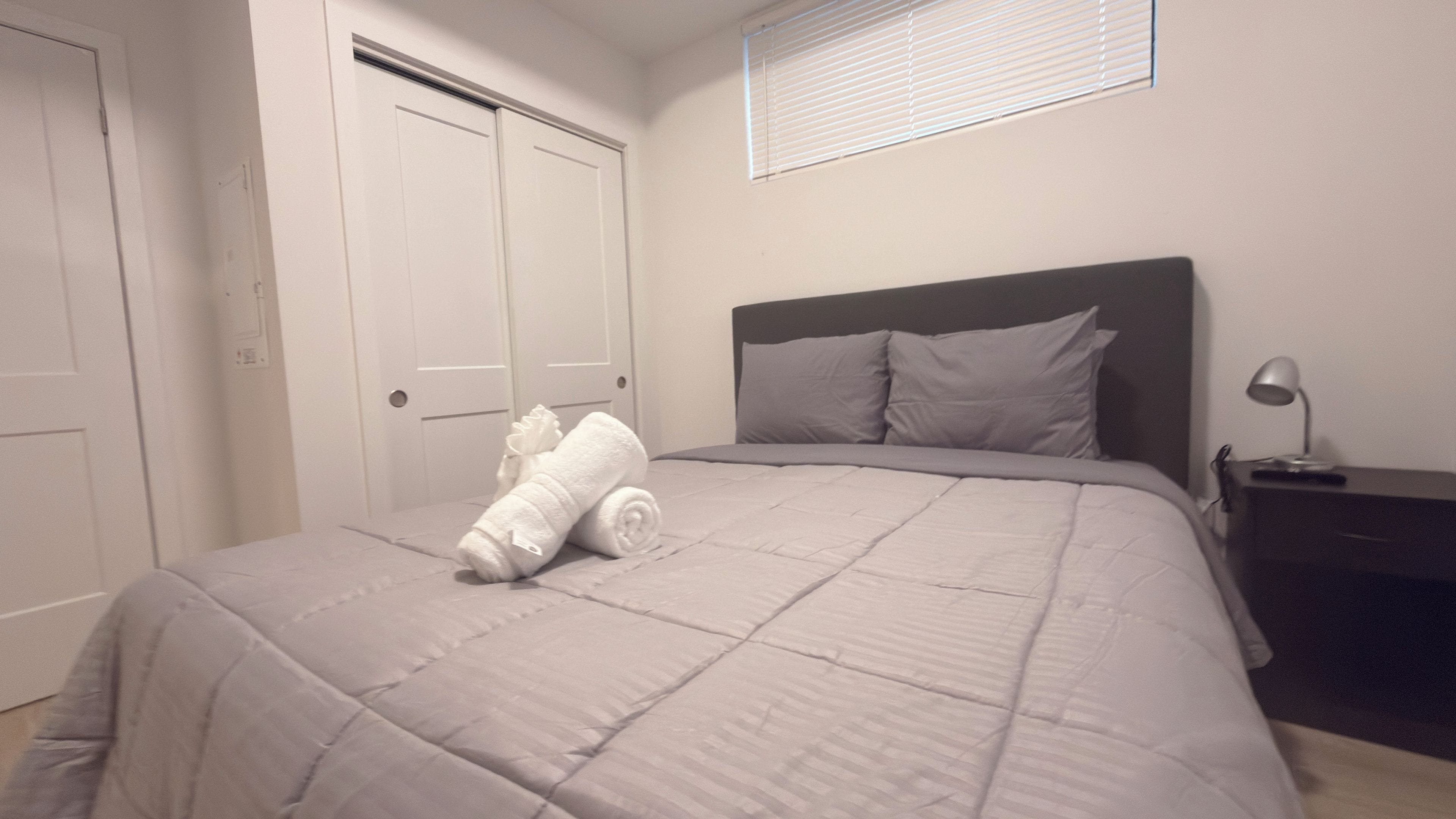 A bedroom with a bed, a nightstand, a lamp, a window with blinds, and a door.