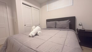 Een slaapkamer met een bed, een nachtkastje, een lamp, een raam met jaloezieën en een deur.