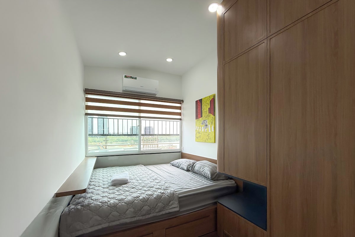 A bedroom with a bed, a window with blinds, a wall-mounted air conditioner, and a wooden wardrobe.