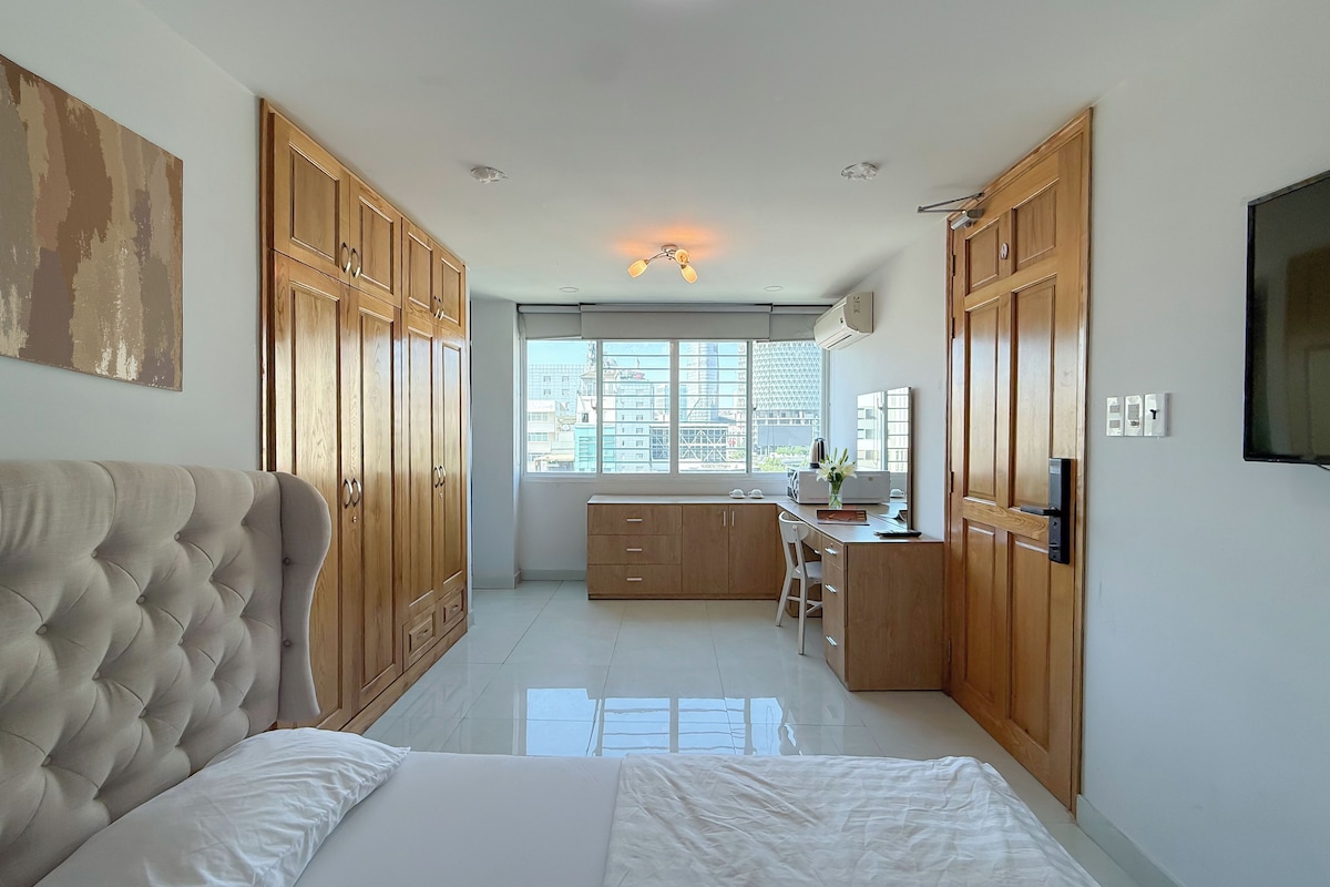 A bedroom with a large bed, wooden wardrobe, and a desk area with a chair and a computer.