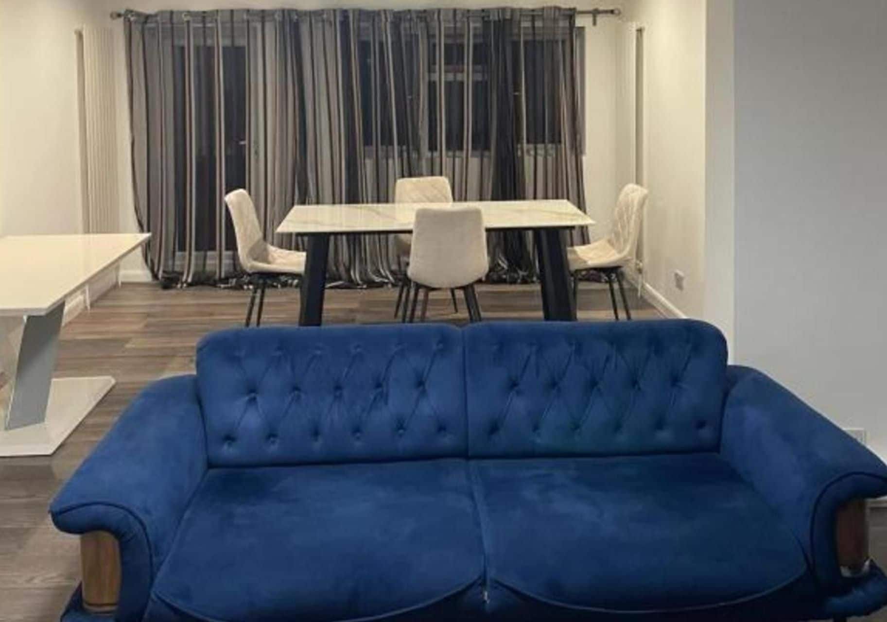 Una habitación con un sofá azul, una mesa de comedor con sillas y una ventana con cortinas.