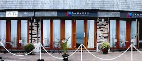 Un edificio con un letrero que dice 'SAWVERS', indicando un restaurante o cafetería. Hay áreas de estar al aire libre con mesas y sillas.