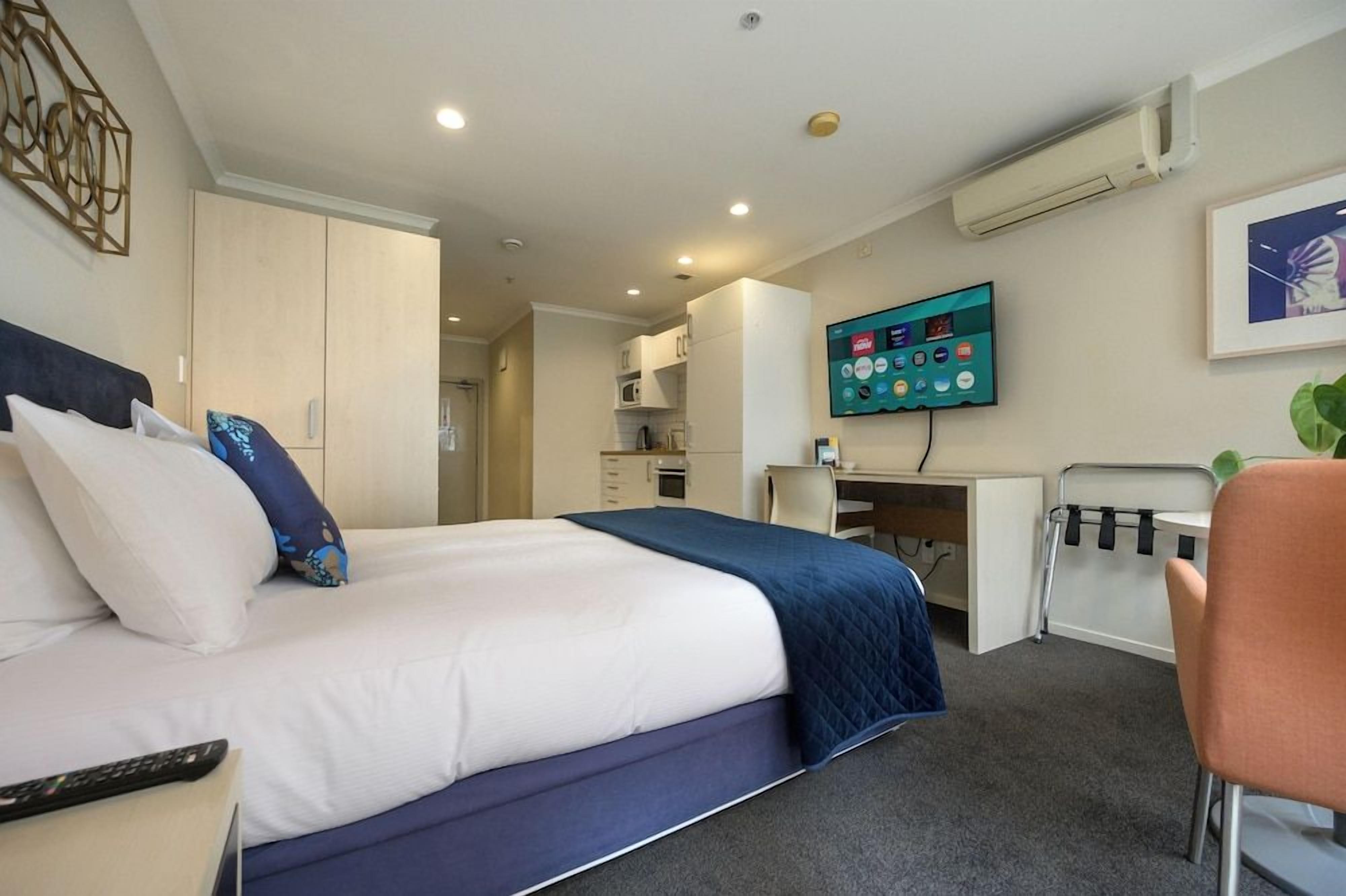 Una habitación de hotel con cama, escritorio, silla, televisor y kitchenette.